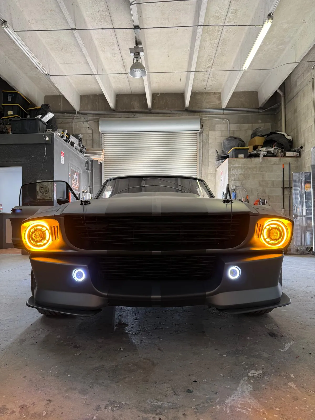 Front view of a custom Mustang fastback with the headlights and fog lights on inside the shop